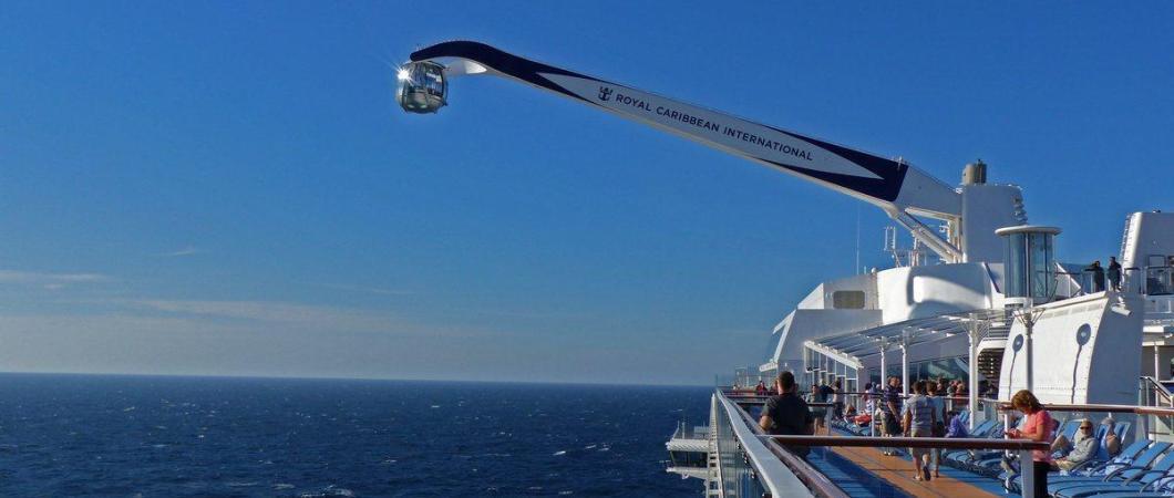 Ball-shaped passenger carying pod on the end of a crane arm dangles out to the side of a cruise ship over blue waters and against a blue sky.