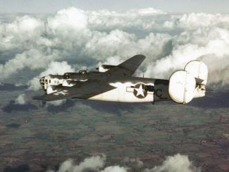 A US Navy four-engined bomber in white & grey flying over fields in England