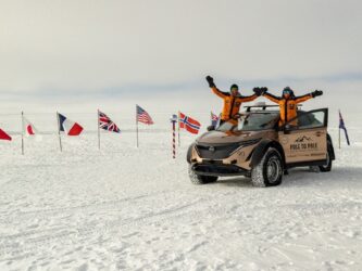 Couple in arctic gear wave from their vehicle in front of national flags at the snow covered South Pole