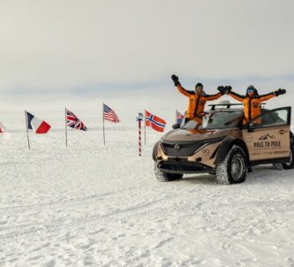Couple in arctic gear wave from their vehicle in front of national flags at the snow covered South Pole