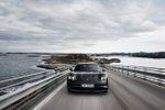 A sports car drives towards the camera on an empty causeway road between rocky islands.