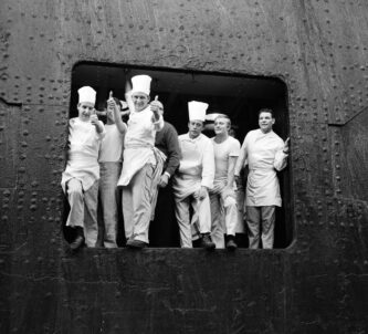 B/W photo of white uniformed chefs and kitchen staff at an open port of a ship