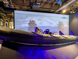 A large black Rigid Inflatable Boat in front of a large screen video showing a RIB in action