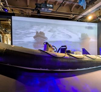 A large black Rigid Inflatable Boat in front of a large screen video showing a RIB in action