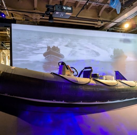 A large black Rigid Inflatable Boat in front of a large screen video showing a RIB in action
