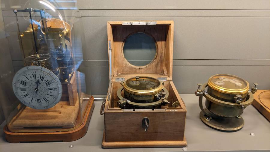 A group of three clocks. The first has a large glass cloche over it. The second is in a wooden box. The third stands alone.