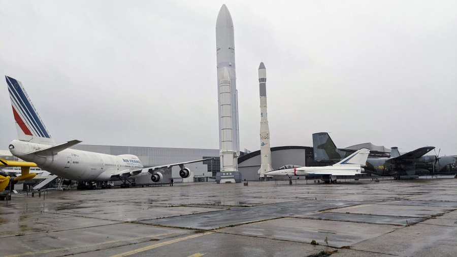View across the tarmac on a rainy day, with the Boeing 474 and two Ariane rockets prominent