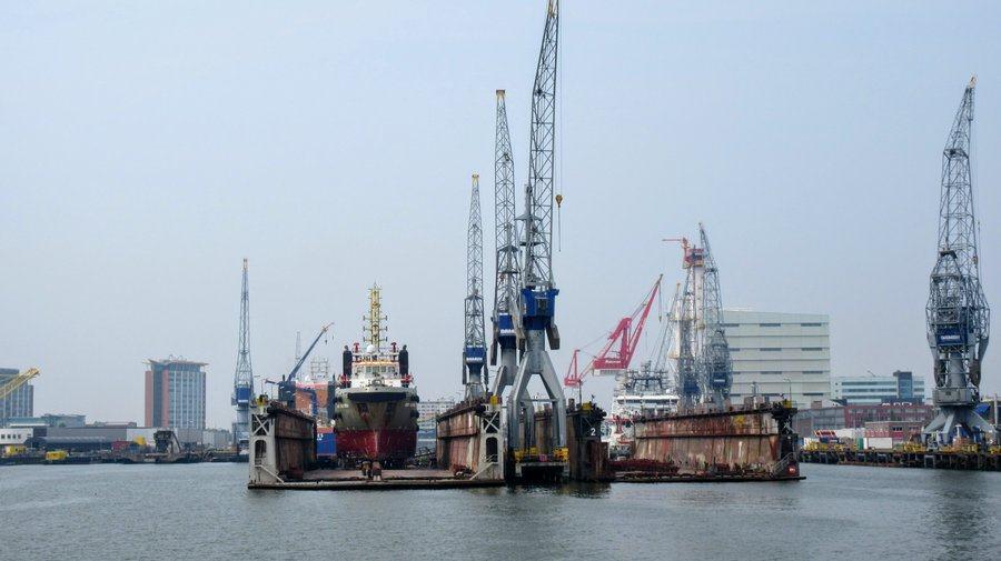 A red bottomed ship out of the water in a floating dry dock with cranes alongside