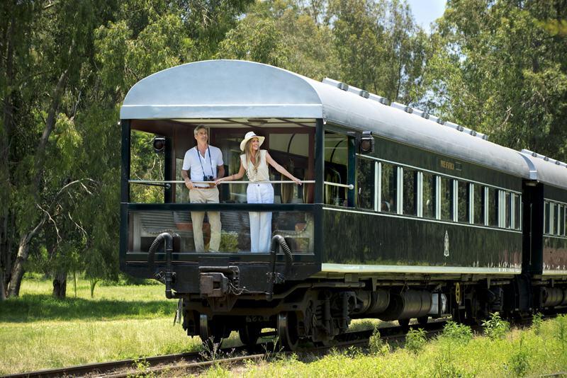 A couple stand looking out from the balcony at the back of a green rail carriage