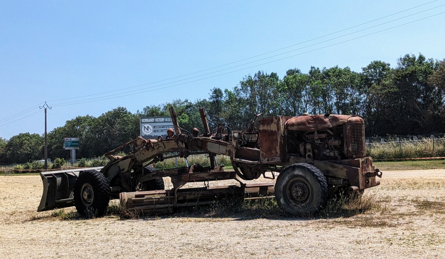 A rusty road grader with a bulldozer blade slung under its arched frame