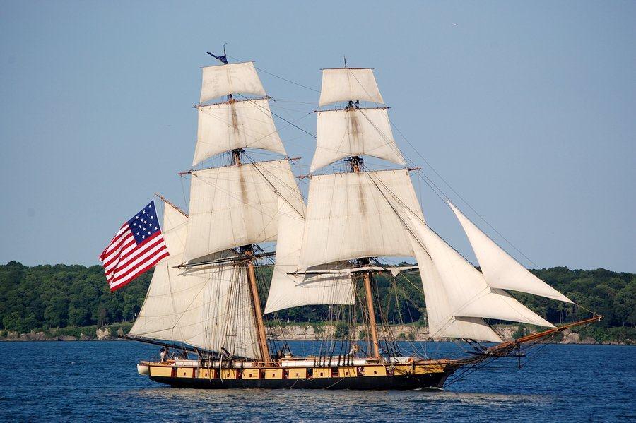 A square rigged sailing ship, the SSV Niagara, sails past a green shoreline. She flies a large Stars & Stripes from her stern.
