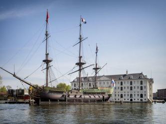 A three masted frigate flying the Dutch flag at her stern is moored in front of the grand classical building of the National Maritime Museum