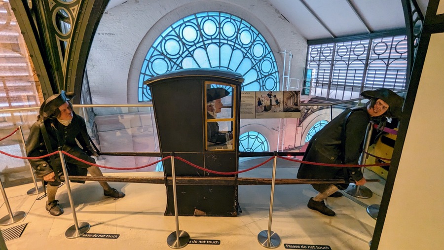 Two men (mannequins) struggling to carry a gentleman in a sedan chair