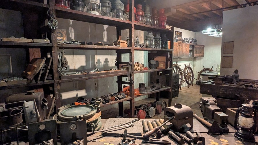 A recreation of a storeroom with a jumble of gear on its shelves and workbenches, including ships' lanterns, machinery, cable tensioners, and portholes