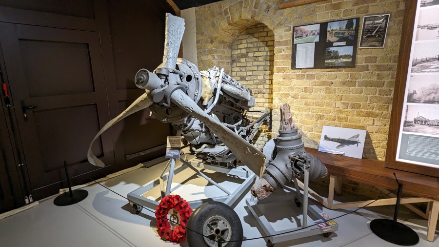 A wrecked light grey aero engine with the stumpy remains of a 3-bladed propeller displayed on an engine stand at the Dynamo Museum, Dunkirk