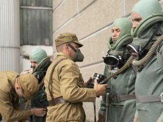 Three men in heavy green suits with masks and breathing apparatus are stood against a wall while two soldiers help to prepare them.