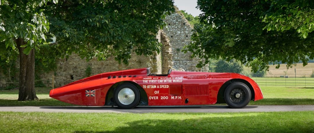 A sleek 100 year old racing car, the Sunbeam 1000hp, in shiny red paint is parked under some trees at the National Motor Museum, Beaulieu