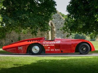 A sleek 100 year old racing car, the Sunbeam 1000hp, in shiny red paint is parked under some trees at the National Motor Museum, Beaulieu