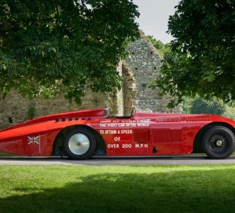 A sleek 100 year old racing car, the Sunbeam 1000hp, in shiny red paint is parked under some trees at the National Motor Museum, Beaulieu