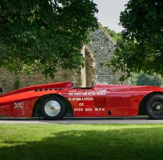 A sleek 100 year old racing car, the Sunbeam 1000hp, in shiny red paint is parked under some trees at the National Motor Museum, Beaulieu
