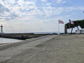 View across an open space to a pier with a lighthouse on it, and at the landward end a stone pillar memorial with two flags flying