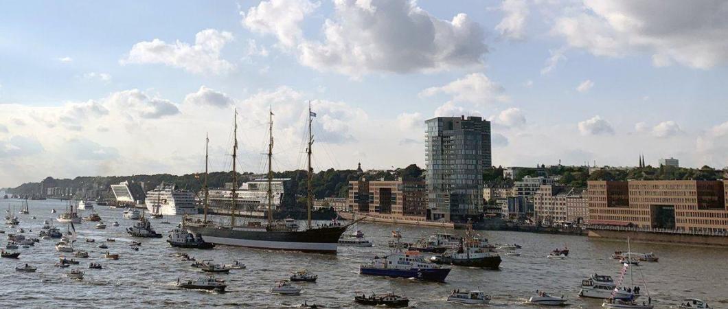 Four-masted barque being towed into port amid a welcome party of dozens of small boats