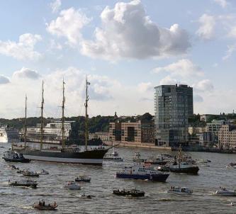 Four-masted barque being towed into port amid a welcome party of dozens of small boats