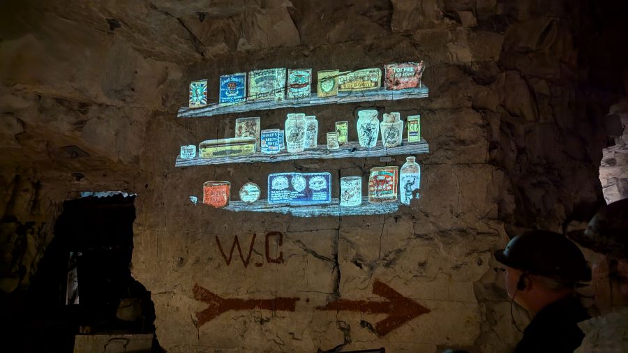 A colour projection on the cavern walls of wooden shelves stocked with food jars & tins
