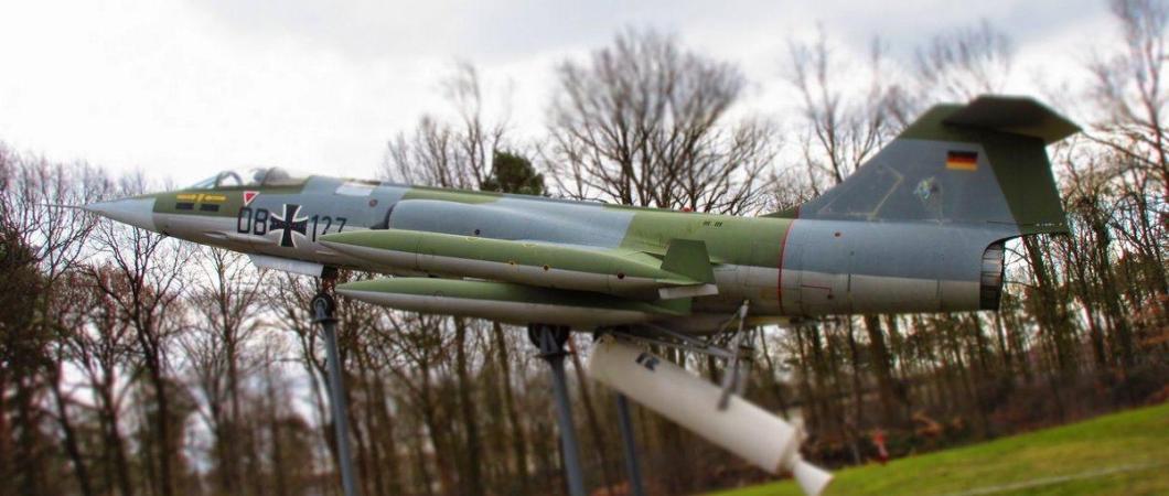 Green and grey pencil-thin German Starfighter with a white JATO rocket underneath, mounted on a display stand outside with grass & tress behind