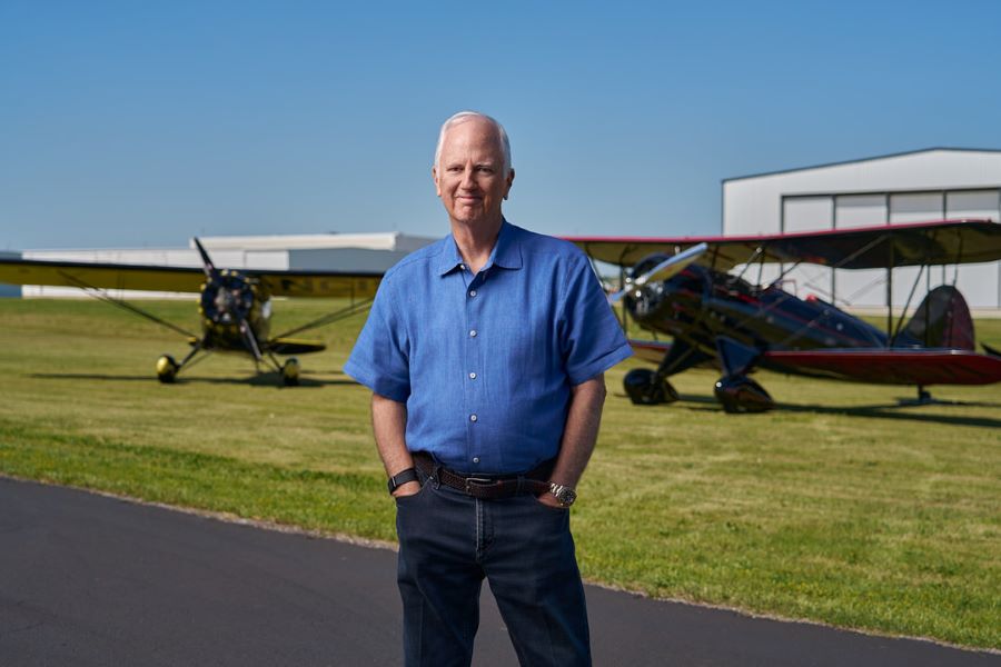 Tony Caldwell, a middle aged man stands in black trousers and a blue shirt in front of two biplanes parked near airport buildings on a sunny day