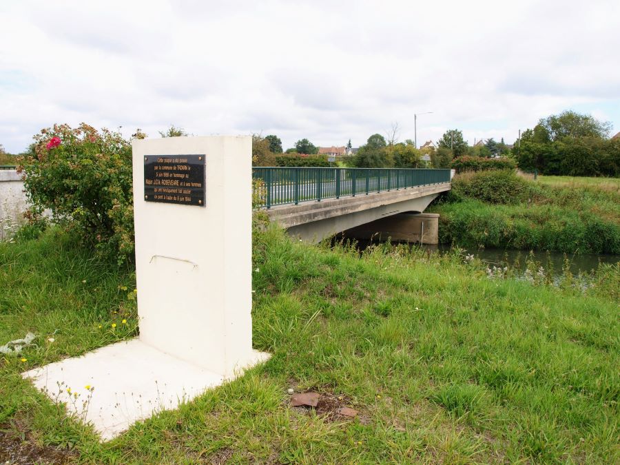 A white stone memorial in the foreground with Troarn bridge behind