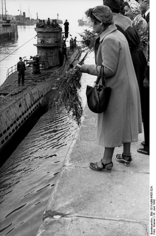 A woman on the quay with other officers and civilians, throws flowers to a U-boat passing below