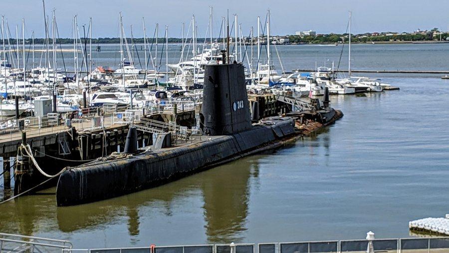 A black submarine berthed alongside a marina with rusty sections of her hull exposed