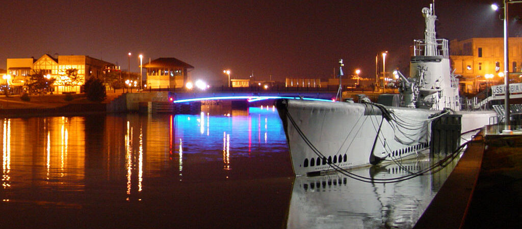 Submarine moored alongside in still water at night with nearby coloured street lights reflecting on the water