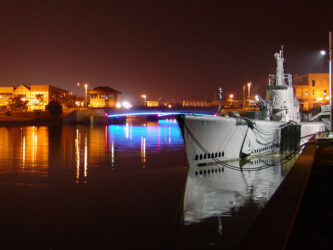 Submarine moored alongside in still water at night with nearby coloured street lights reflecting on the water