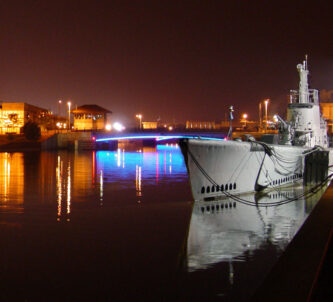 Submarine moored alongside in still water at night with nearby coloured street lights reflecting on the water