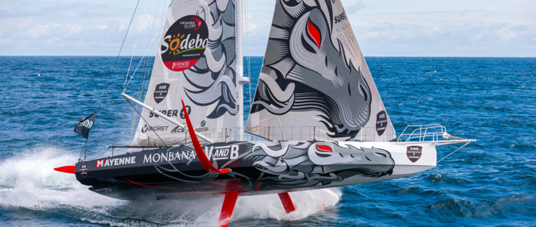 An IMOCA racing yacht rises dramatically above the sea on its foils