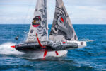 An IMOCA racing yacht rises dramatically above the sea on its foils