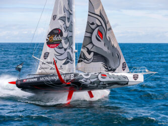 An IMOCA racing yacht rises dramatically above the sea on its foils