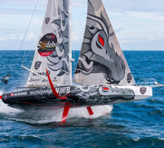 An IMOCA racing yacht rises dramatically above the sea on its foils