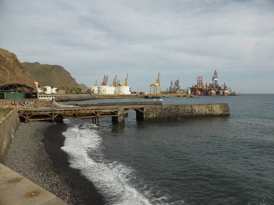 A small stony grey beach with industrial fuel tanks and docks behind