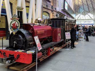 A small maroon coloured steam engine on temporary tracks in the passenger concourse