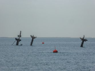 Three T-shaped masts stick out of the sea with a red warning buoy in the foreground