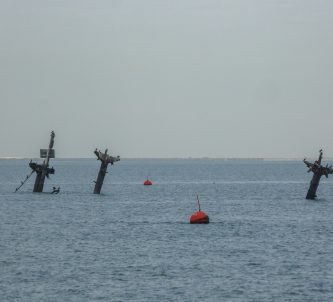 Three T-shaped masts stick out of the sea with a red warning buoy in the foreground
