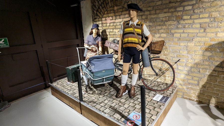 Dynamo Museum diorama: A woman and a young boy (mannequins) on a cobbled street with suitcases, a pram, and a bicycle